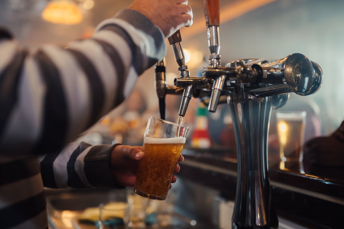 Bartender's hand pouring a fresh pint of golden craft beer from a tap, filling a glass in a dimly lit bar setting