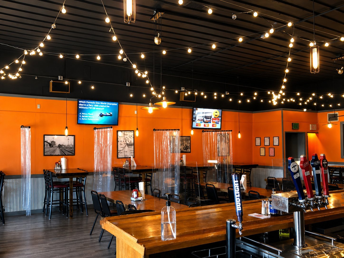 Modern brewery or bar interior with bright orange walls, wooden tables, string lights, and TV screens mounted on walls